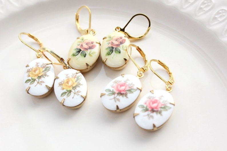 May include: Four pairs of gold tone earrings with oval shaped porcelain charms featuring floral designs. The charms have a white background with pink, yellow, and green floral patterns.