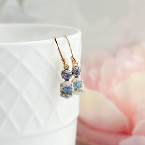 Puede incluir: Un par de pendientes de oro con un dise&ntilde;o floral azul y blanco y un acento de cristal morado.