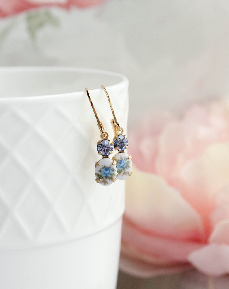 Puede incluir: Un par de pendientes de oro con un dise&ntilde;o floral azul y blanco y un acento de cristal morado.