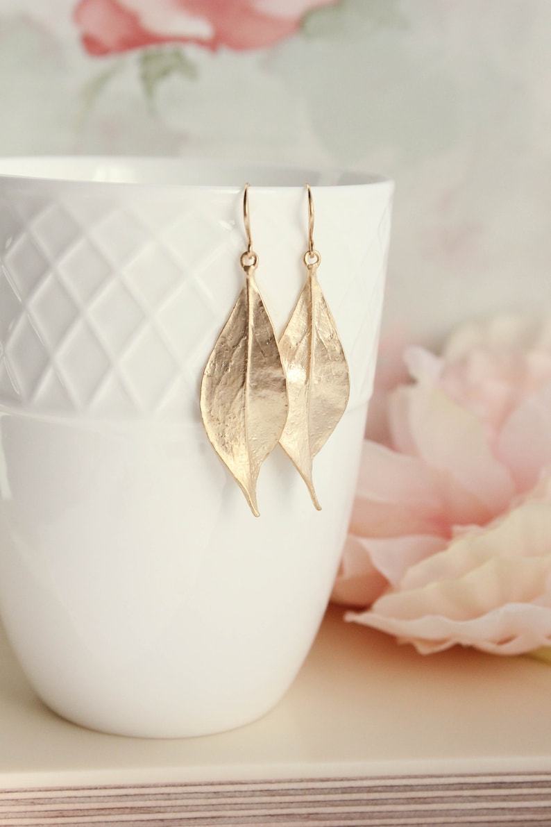 K&ouml;nnte beinhalten: Goldene Blatt-Ohrringe mit Textur. Die Ohrringe h&auml;ngen an einer wei&szlig;en Tasse.