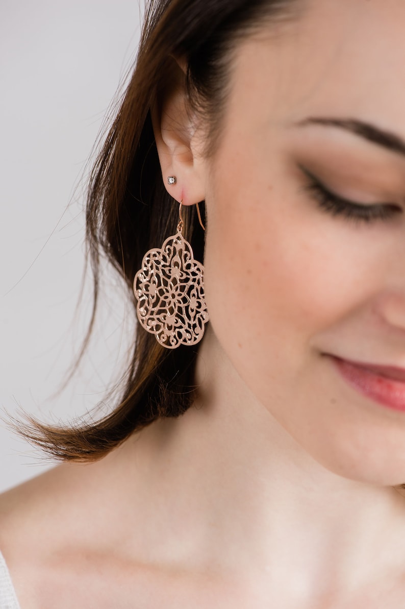 May include: Rose gold filigree earrings with a delicate, intricate design. The earrings are dangle style and feature a circular shape.