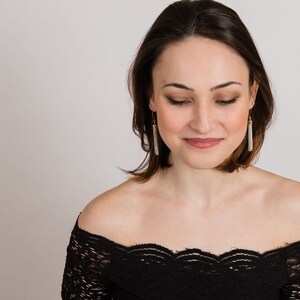 May include: A woman with short brown hair wearing a black off-the-shoulder lace top and long silver tassel earrings.