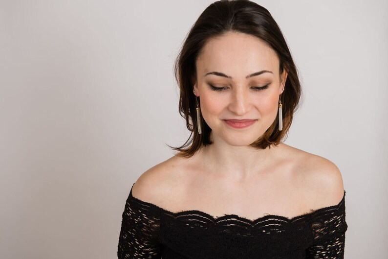 May include: A woman with short brown hair wearing a black off-the-shoulder lace top and long silver tassel earrings.