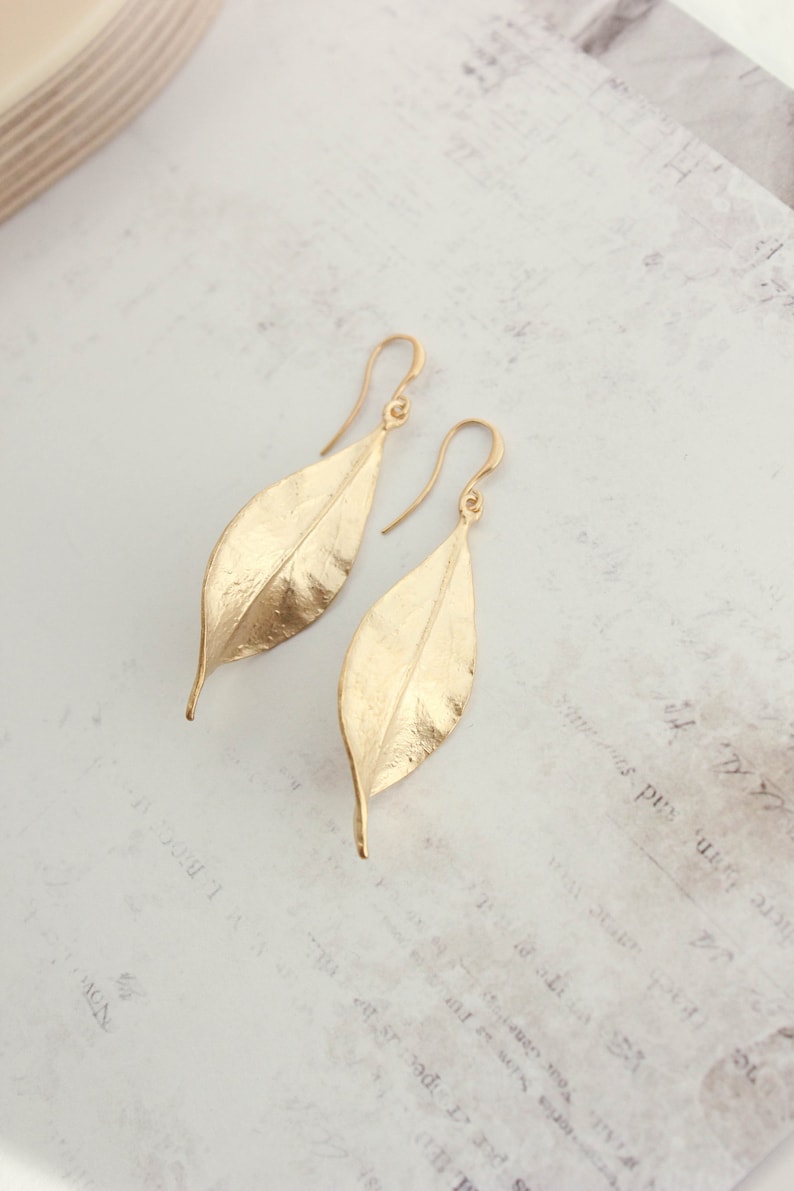 K&ouml;nnte beinhalten: Ein Paar goldene Ohrringe in Blattform. Die Ohrringe sind aus strukturiertem Metall gefertigt und haben ein schlichtes, elegantes Design.