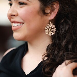 May include: A woman with long, curly brown hair wearing gold dangle earrings with a delicate, intricate design.