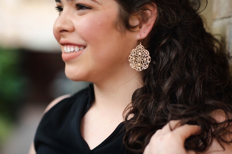 May include: A woman with long, curly brown hair wearing gold dangle earrings with a delicate, intricate design.