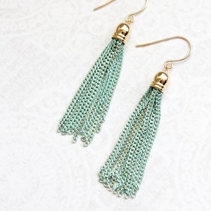 May include: A pair of long, dangling earrings with a light blue tassel made of chain links. The earrings have gold hooks and a gold bead at the top of each tassel.
