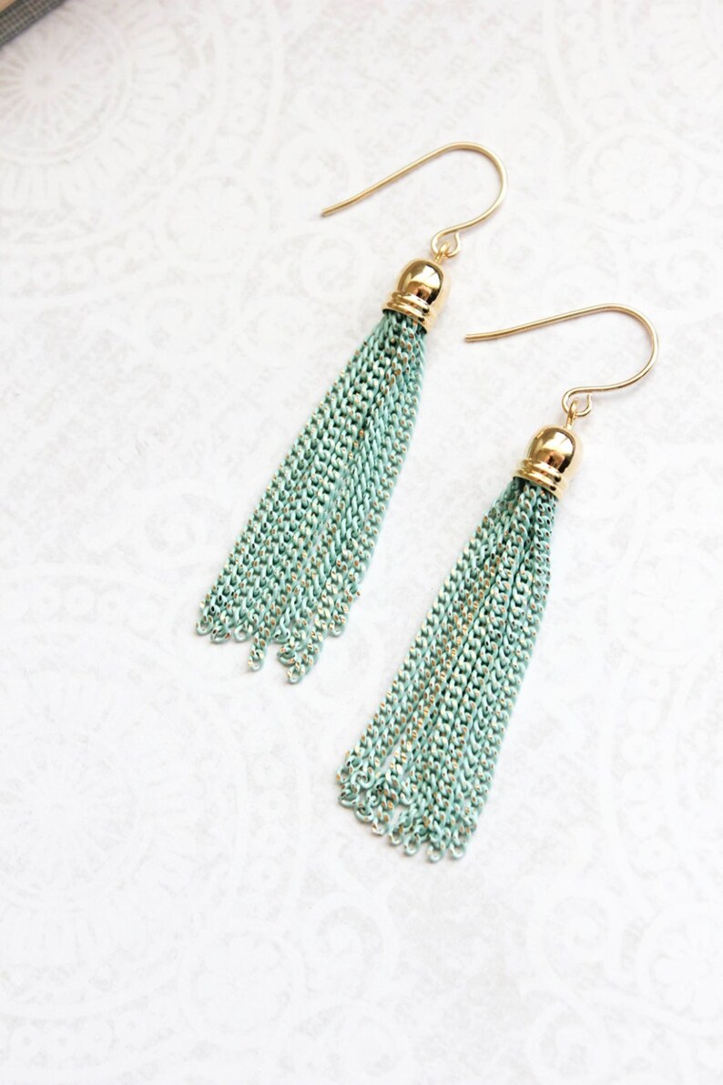 May include: A pair of long, dangling earrings with a light blue tassel made of chain links. The earrings have gold hooks and a gold bead at the top of each tassel.