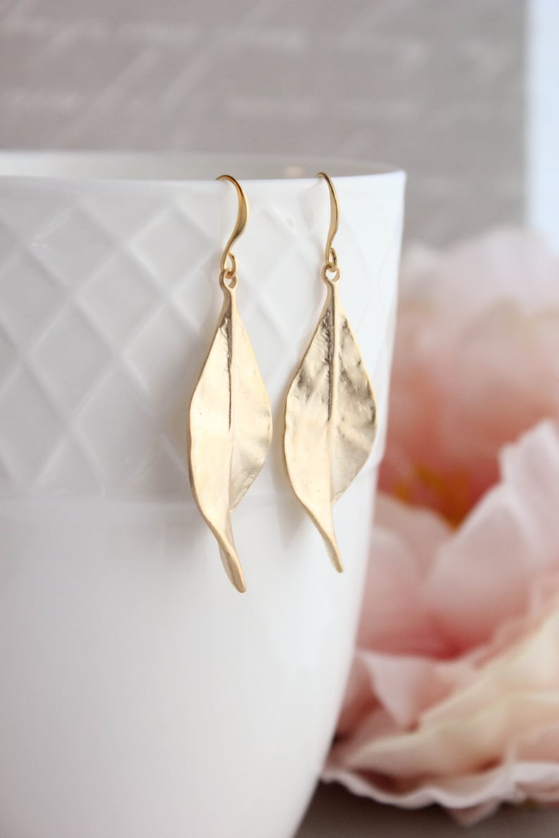 K&ouml;nnte beinhalten: Ein Paar goldene Ohrringe in Blattform. Die Ohrringe sind aus strukturiertem Metall gefertigt und haben ein schlichtes, elegantes Design.