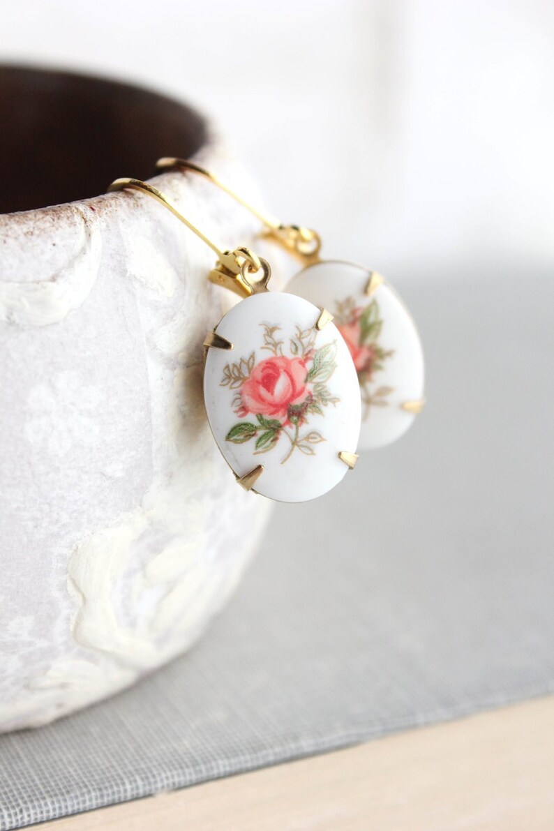 May include: A pair of gold earrings with oval-shaped porcelain charms featuring a pink rose floral design.