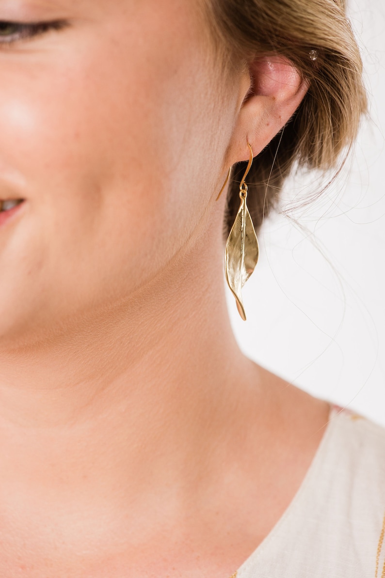 K&ouml;nnte beinhalten: Goldene, tropfenf&ouml;rmige Ohrringe mit Blattform und geh&auml;mmerter Textur. Die Ohrringe werden von einer Person mit blonden Haaren getragen.