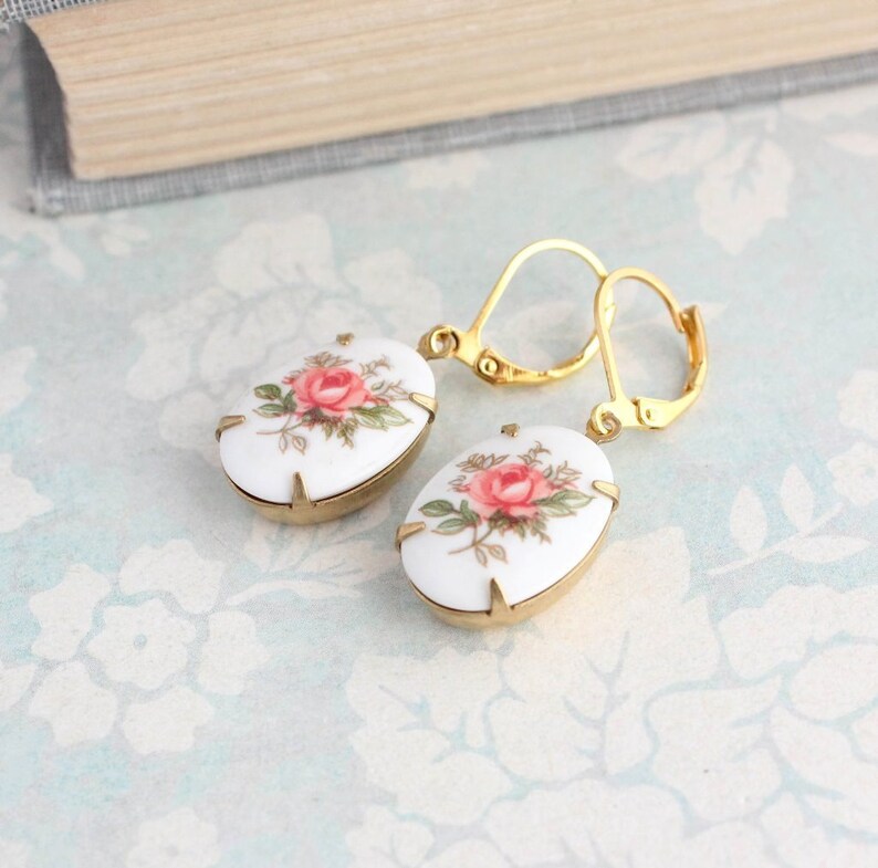 May include: A pair of gold earrings with white porcelain oval-shaped charms featuring pink and green floral designs.
