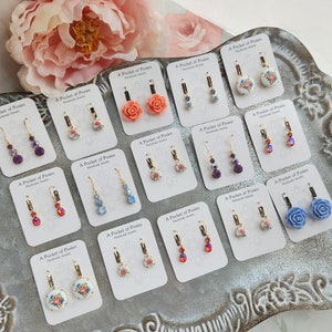 Puede incluir: Surtido de pendientes hechos a mano con dise&ntilde;os florales y detalles de cristal. Los pendientes se muestran en una bandeja plateada con fondo blanco. Cada pendiente est&aacute; etiquetado con "A Pocket of Posies Handmade Jewelry".