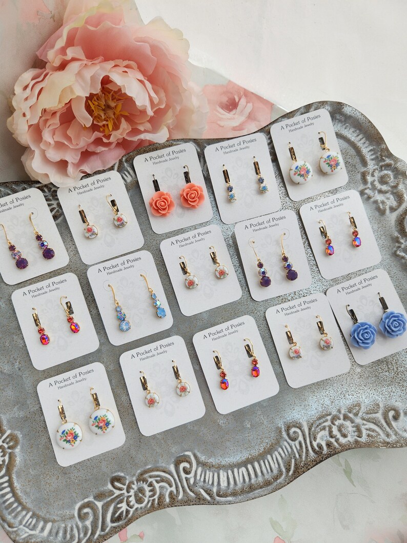 Puede incluir: Surtido de pendientes hechos a mano con dise&ntilde;os florales y detalles de cristal. Los pendientes se muestran en una bandeja plateada con fondo blanco. Cada pendiente est&aacute; etiquetado con "A Pocket of Posies Handmade Jewelry".