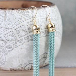 May include: A pair of gold and light blue tassel earrings. The earrings are made of a chain-like material and have a gold ball at the top.
