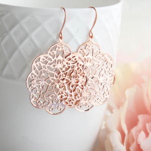 May include: Rose gold dangle earrings with a delicate, intricate, circular, floral design.