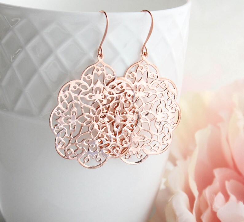 May include: Rose gold dangle earrings with a delicate, intricate, circular, floral design.