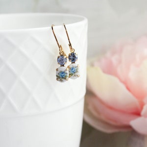 Puede incluir: Un par de pendientes de oro con un dise&ntilde;o floral azul y un peque&ntilde;o cristal azul. Los pendientes est&aacute;n colgando de una taza blanca.