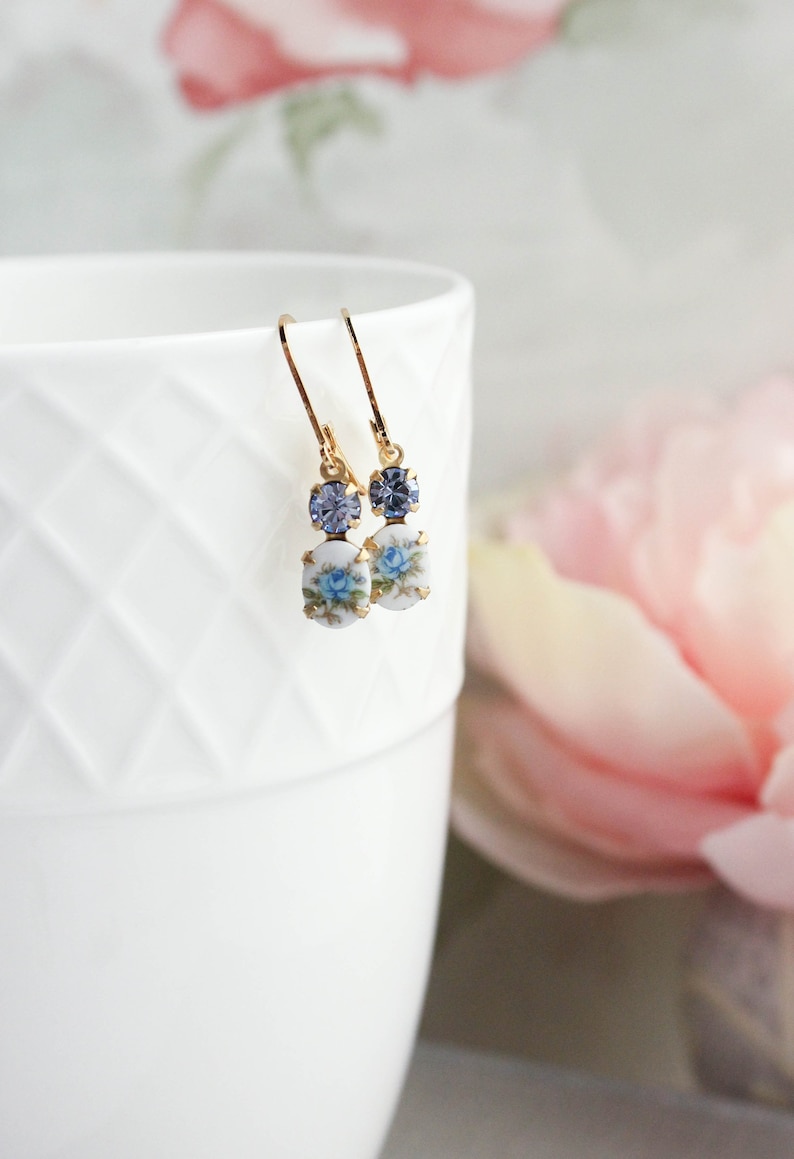 Puede incluir: Un par de pendientes de oro con un dise&ntilde;o floral azul y un peque&ntilde;o cristal azul. Los pendientes est&aacute;n colgando de una taza blanca.