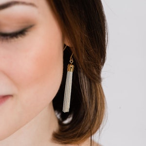 May include: A close-up of a woman's ear with a gold and silver tassel earring. The earring is made of a chain-like material and hangs down from the earlobe.