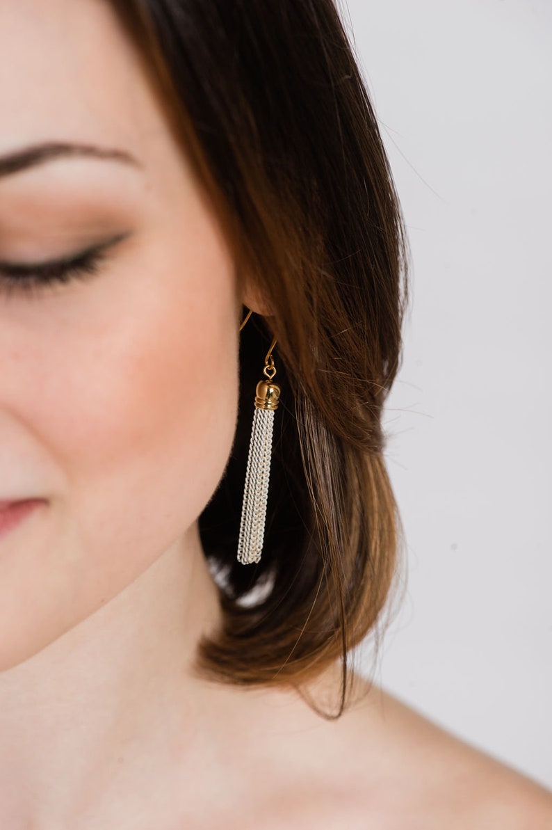 May include: A close-up of a woman's ear with a gold and silver tassel earring. The earring is made of a chain-like material and hangs down from the earlobe.