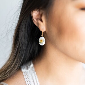 May include: A pair of gold earrings with a white oval pendant featuring a yellow rose and green leaves.