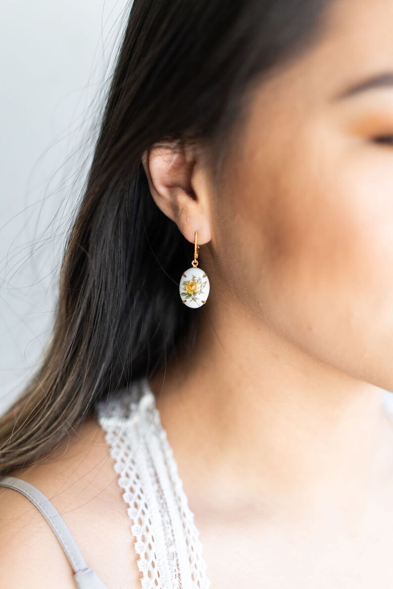 May include: A pair of gold earrings with a white oval pendant featuring a yellow rose and green leaves.