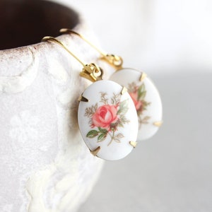 May include: A pair of gold tone earrings with oval-shaped white porcelain charms featuring a pink rose floral design.