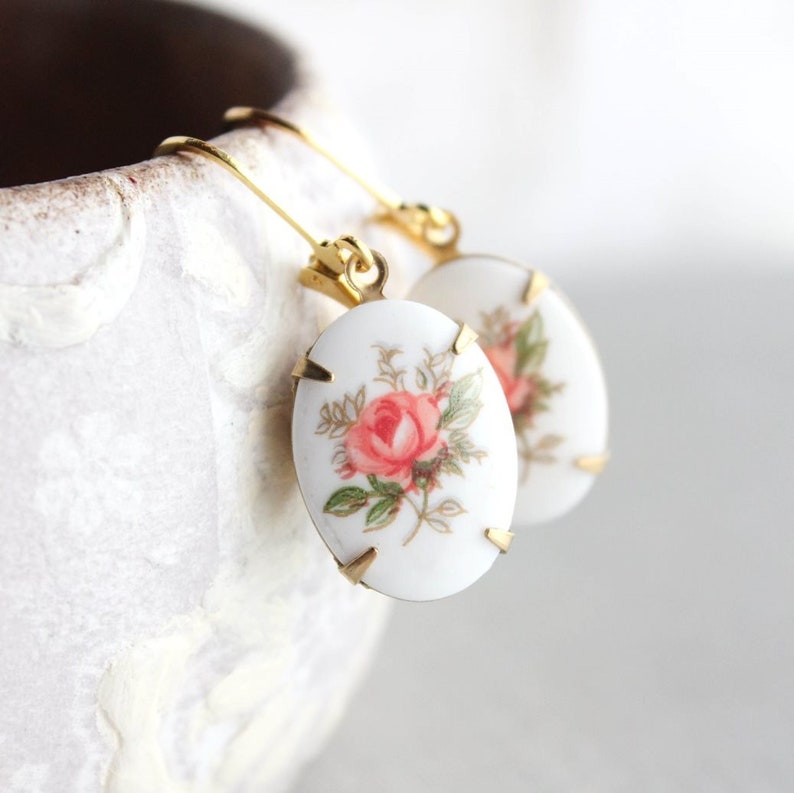 May include: A pair of gold tone earrings with oval-shaped white porcelain charms featuring a pink rose floral design.