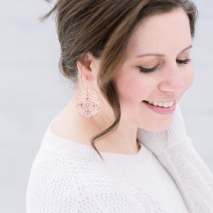 May include: A woman with brown hair wearing a white sweater and rose gold earrings. The earrings are a delicate, intricate design.