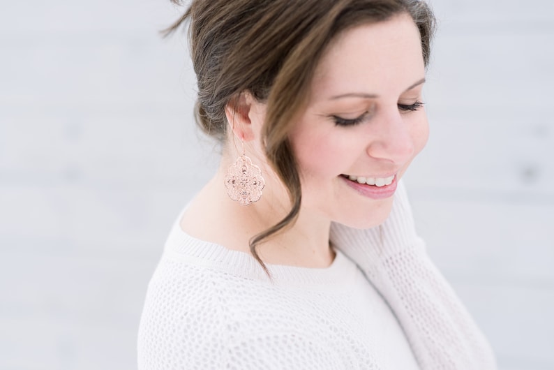 May include: A woman with brown hair wearing a white sweater and rose gold earrings. The earrings are a delicate, intricate design.
