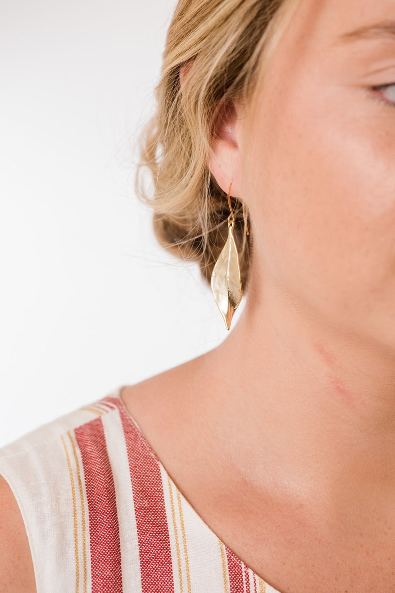 K&ouml;nnte beinhalten: Goldene Blatt-Ohrringe mit H&auml;mmerung. Die Ohrringe werden von einer Person mit blonden Haaren und einem rot-wei&szlig; gestreiften Shirt getragen.
