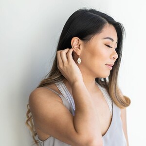 May include: A woman with long brown hair wearing a gray tank top and gold earrings. She is looking down and touching her ear.