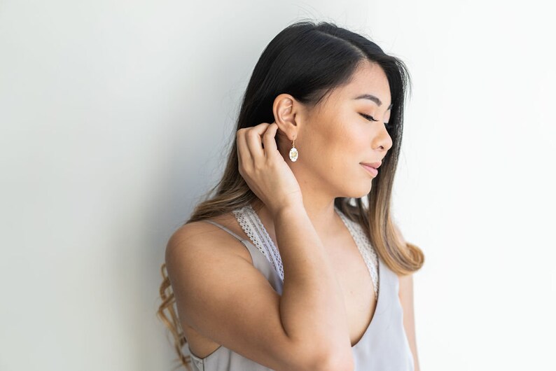 May include: A woman with long brown hair wearing a gray tank top and gold earrings. She is looking down and touching her ear.