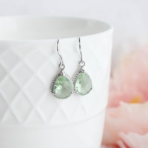 Puede incluir: Un par de pendientes de plata con piedras preciosas de forma de pera de color verde claro. Los pendientes tienen un dise&ntilde;o delicado e intrincado.