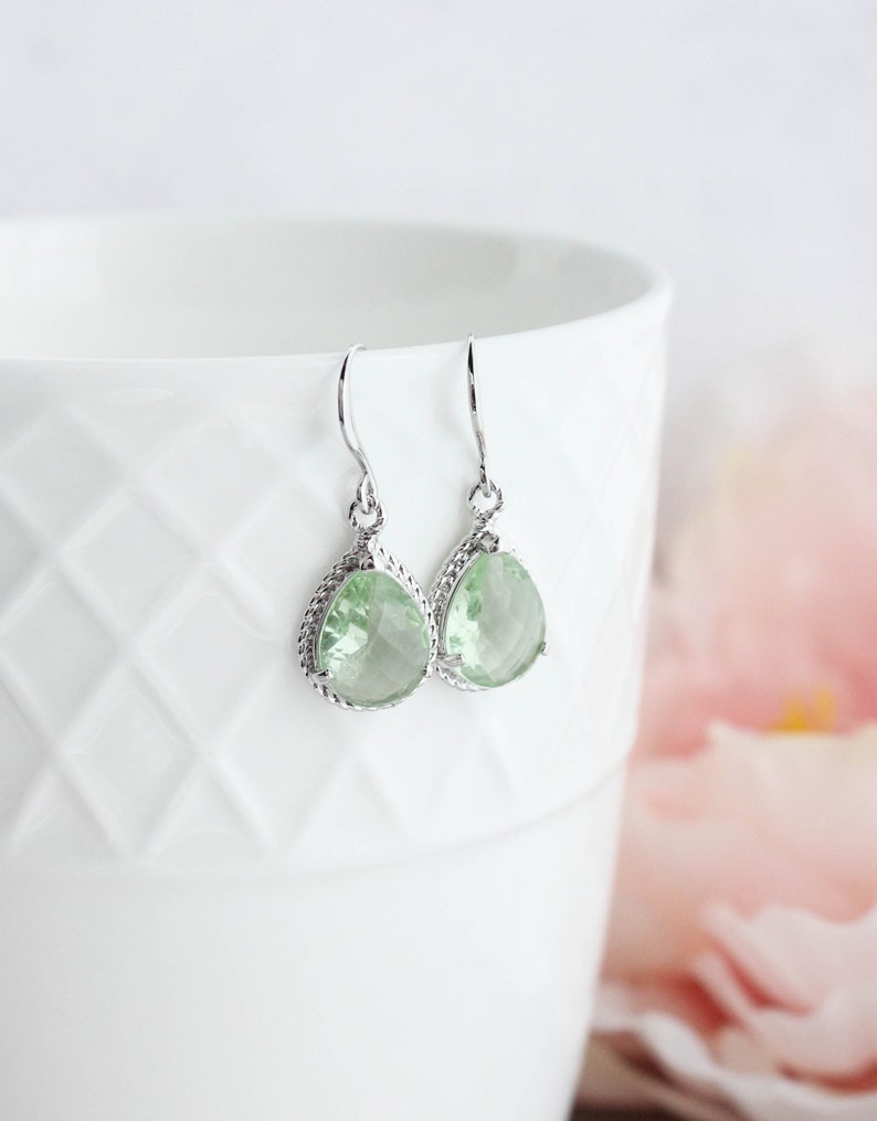 Puede incluir: Un par de pendientes de plata con piedras preciosas de forma de pera de color verde claro. Los pendientes tienen un dise&ntilde;o delicado e intrincado.