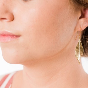 K&ouml;nnte beinhalten: Goldene Blatt-Ohrringe mit einer texturierten Oberfl&auml;che. Die Ohrringe werden von einer Person mit blonden Haaren getragen.