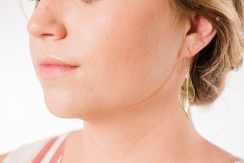 K&ouml;nnte beinhalten: Goldene Blatt-Ohrringe mit einer texturierten Oberfl&auml;che. Die Ohrringe werden von einer Person mit blonden Haaren getragen.