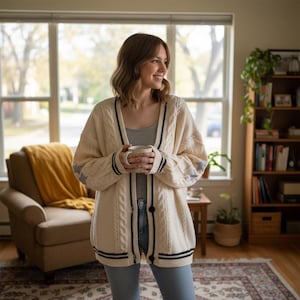 May include: Cream-colored cable knit cardigan with black and navy blue stripes. The cardigan has a V-neck, button closure, and star accents on the sleeves. The person is holding a white mug.