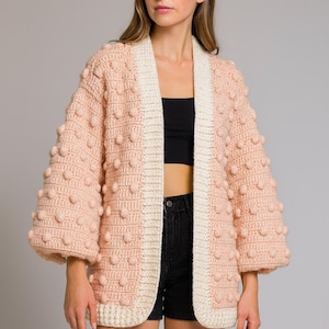 May include: A pale pink, open-front cardigan with a cream trim and bobble detailing. The sleeves are three-quarter length. The cardigan is made of a chunky knit material. The model is wearing a black top and black shorts.