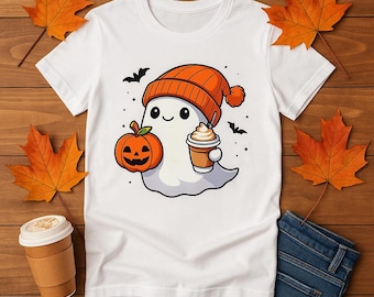 May include: White t-shirt featuring a cartoon ghost wearing an orange beanie and holding a pumpkin and a coffee cup. The ghost has a friendly expression. Black bats and orange leaves decorate the background. The shirt is on a wooden surface.