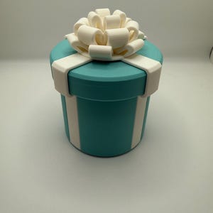 May include: A cylindrical gift box in a vibrant turquoise color featuring a white lid and a white ribbon design that wraps around the sides. A large, three-dimensional white bow sits prominently on top of the lid, completing the gift box appearance.