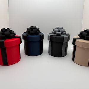 May include: Four cylindrical gift boxes in red, navy blue, black, and tan, each with a matching ribbon and a black bow on top. The boxes are arranged in a row against a white background, showcasing a variety of colors and styles.