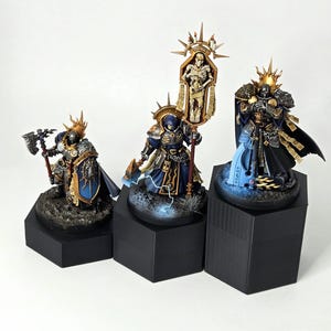 May include: Three miniature figurines of armored characters, each standing on a black hexagonal base. The figures are painted with gold, blue, and black details, and feature ornate helmets and weapons. One figure holds a banner with a skeletal figure.