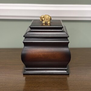 Könnte beinhalten: Eine dunkelbraune Holzkiste mit einer goldenen Elefantenfigur auf der Oberseite. Die Kiste hat eine rechteckige Form mit einem geschwungenen unteren Teil und einem abgestuften Deckel. Der Elefant ist klein und detailliert.