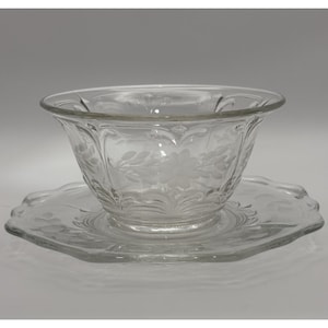 May include: A clear glass bowl and saucer set. The bowl has a flared rim and is decorated with etched floral patterns. The saucer is hexagonal with a scalloped edge and also features etched floral designs. The set is made of clear glass.