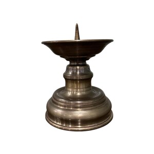 Pode incluir: Um castiçal de cor bronze com uma tigela larga e rasa e um espinho central. A base é redonda e em camadas, com um acabamento liso e polido. O castiçal foi projetado para segurar uma única vela.