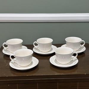 Può includere: Cinque tazze da tè bianche e piattini sono disposte su una superficie di legno scuro. Ogni tazza ha un manico curvo e poggia su un piattino abbinato. Le tazze e i piattini sono realizzati in un materiale bianco lucido.