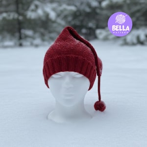 May include: A knitted burgundy hat with a long, pointed top and a small pom-pom. The hat is displayed on a white mannequin head in a snowy outdoor setting. The hat is a winter accessory.