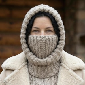 May include: A woman wearing a beige knitted hood and face covering, with a matching sweater and coat. The hood and face covering are made of a thick, ribbed knit material. The coat is a light beige color with a fuzzy lining.
